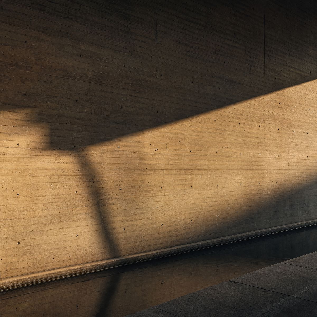 Board-formed concrete wall crossed by a blade of warm sunlight above a still black water channel